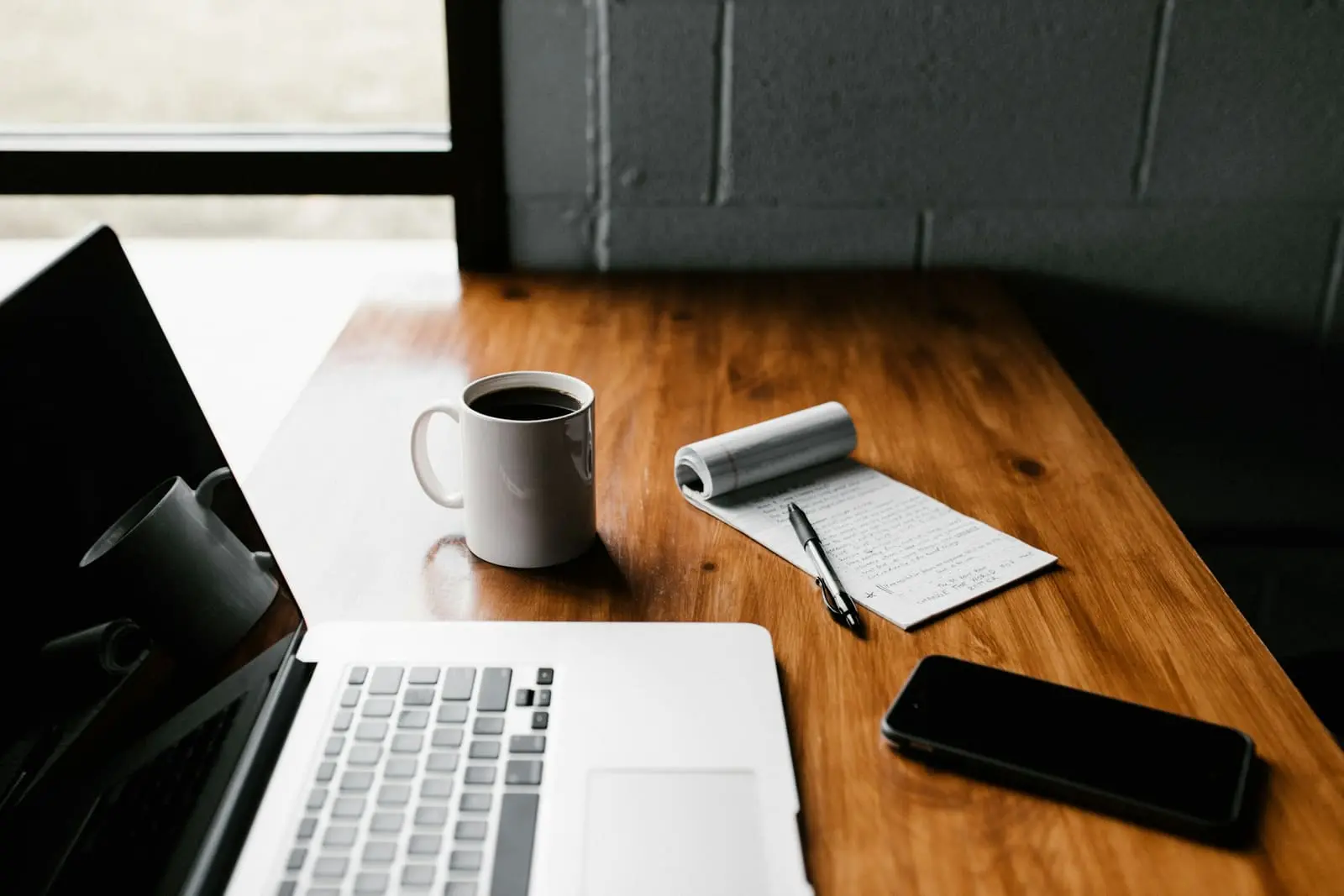 A laptop, a mug, a phone and a notebook on a wooden desk by a window